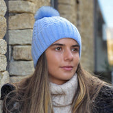 Woman wearing light blue recycled yarn herringbone cable knit hat with pom pom outdoors